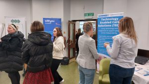A room of people with a woman talking to a person in front of a Technology Enabled Care banner.